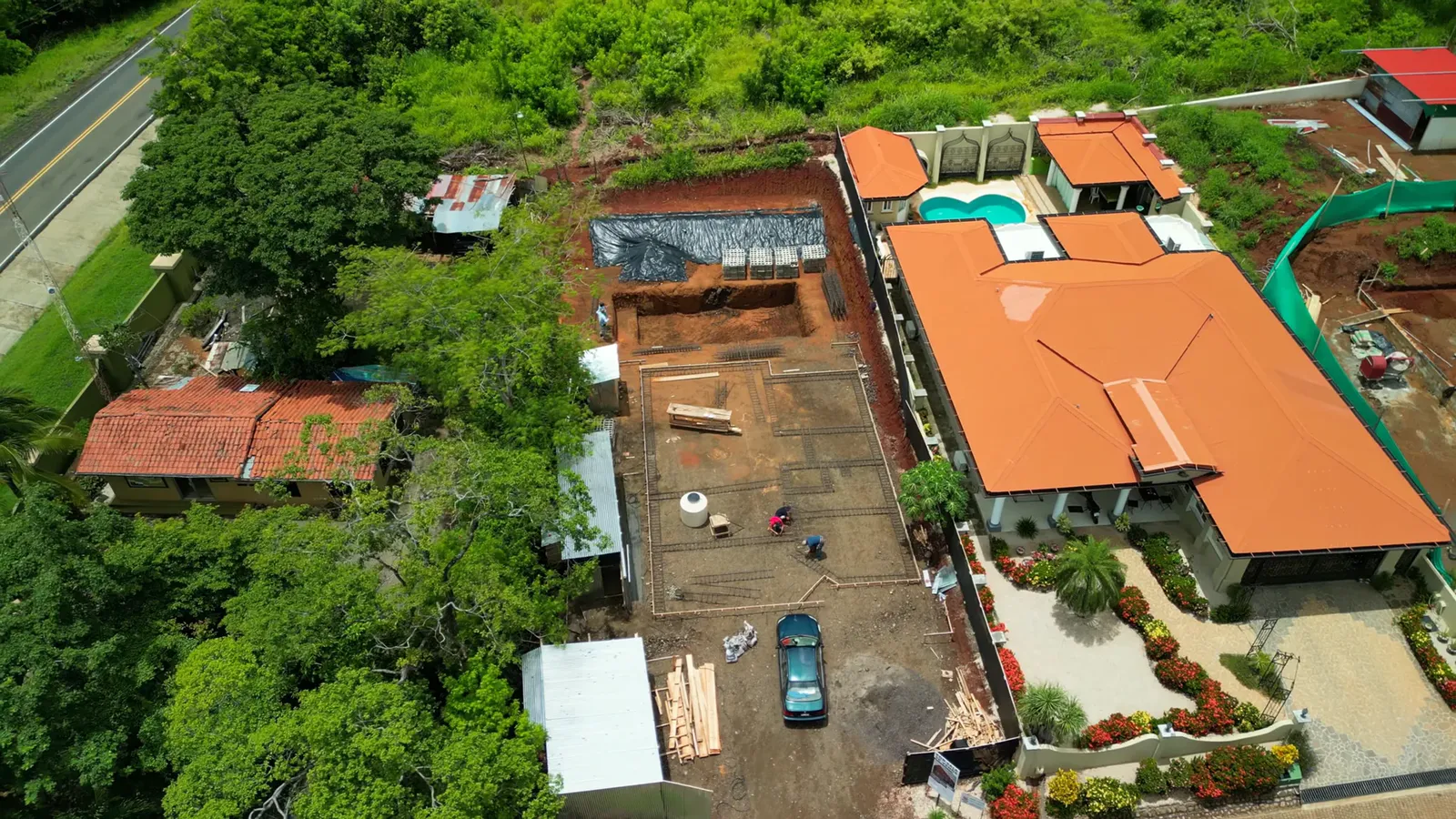 Top-down drone shot of a lot being built beside a finished house with a pool and garden